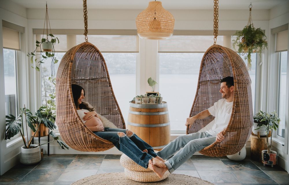Two woven rattan hanging egg chairs flank a wooden barrel in a bright sunlit room with houseplants.
