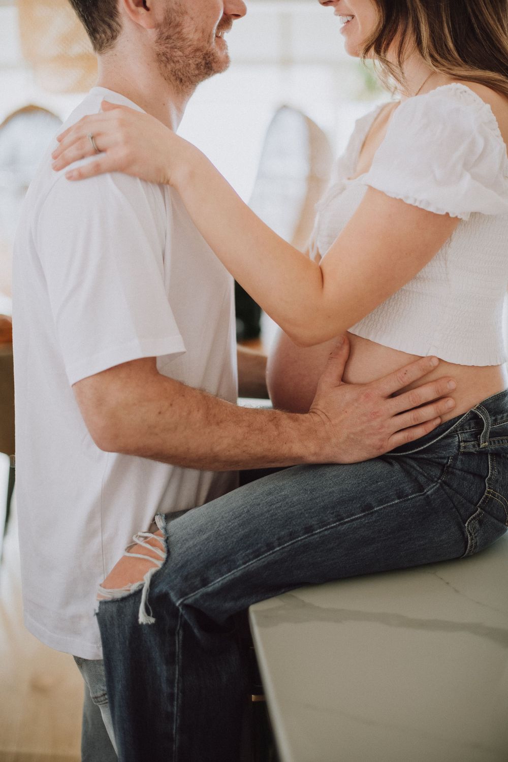 Intimate couple in casual wear embracing indoors, with woman sitting on counter wearing white top and jeans.