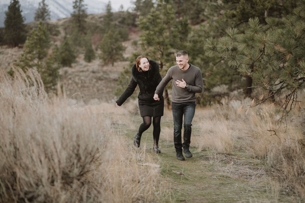 Two people run playfully along a trail through dry brush and evergreen trees on a cloudy winter day.