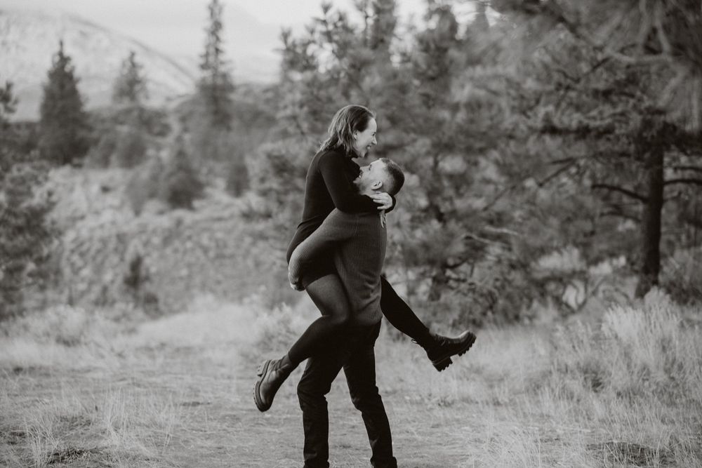 A couple embraces joyfully on a mountain trail surrounded by pine trees in black and white.