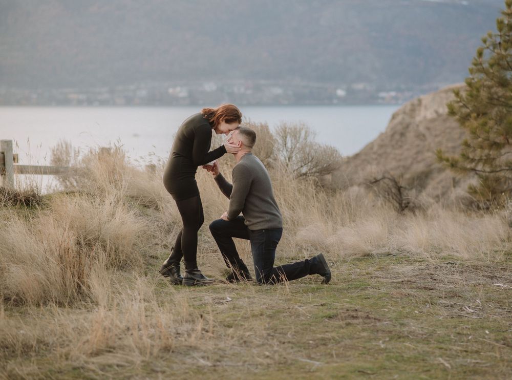 A romantic proposal moment captured on a scenic cliff overlooking a lake with dried grass and mountains in the background.