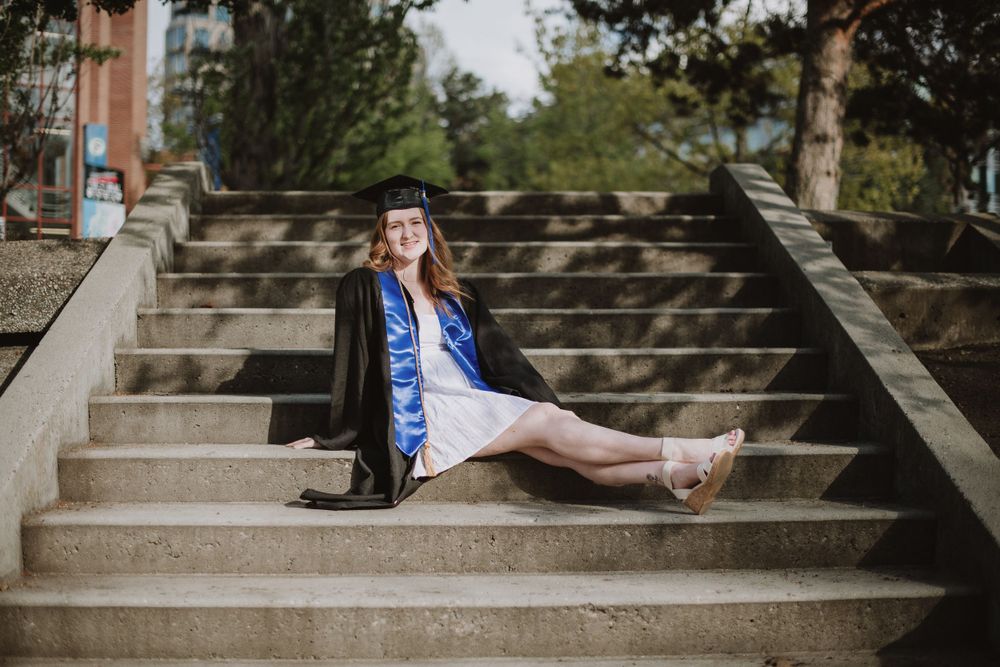 Graduate in cap and gown poses elegantly on outdoor concrete steps surrounded by trees.