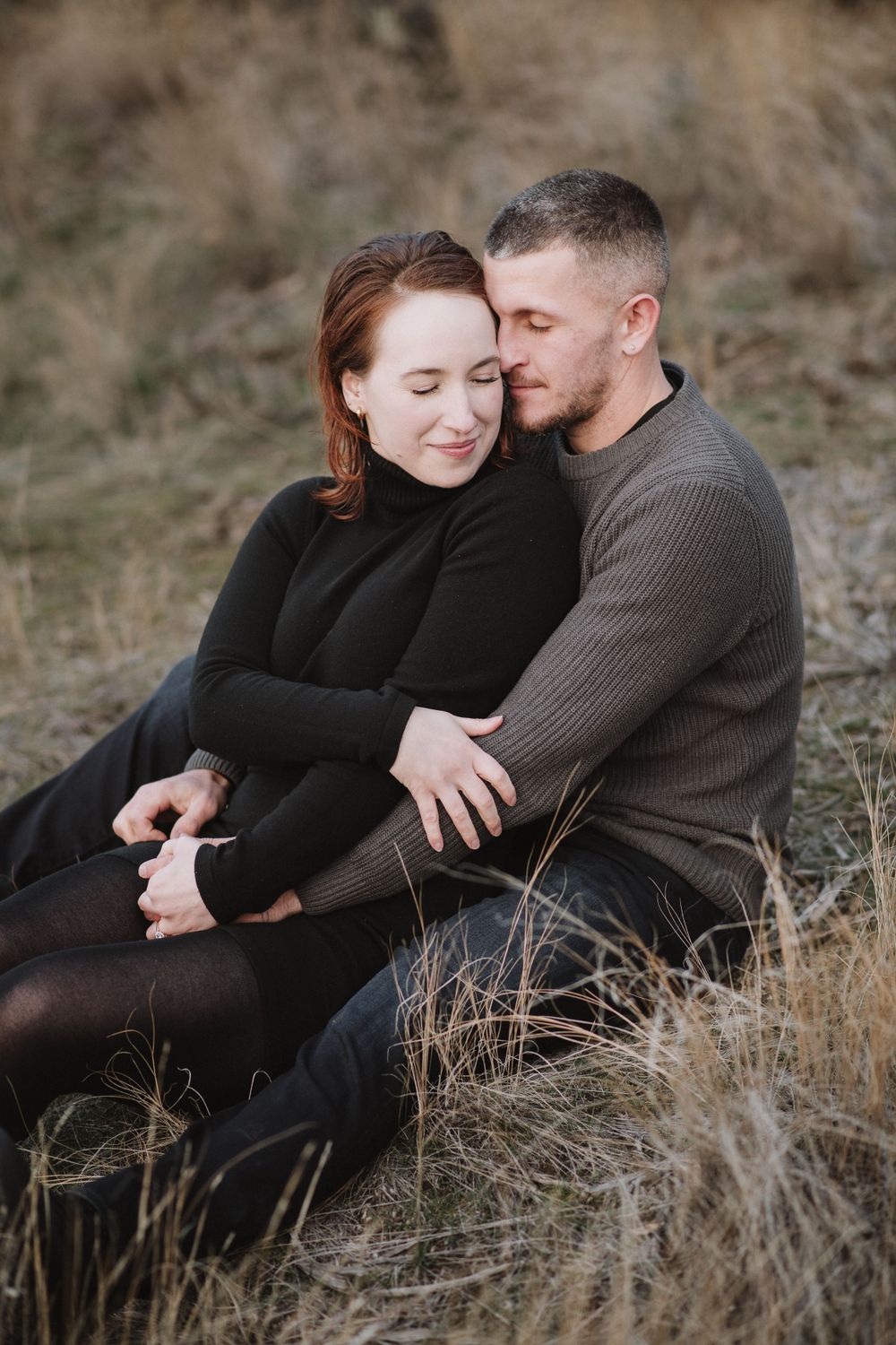 A couple in cozy sweaters share an intimate moment while sitting together in tall dry grass.