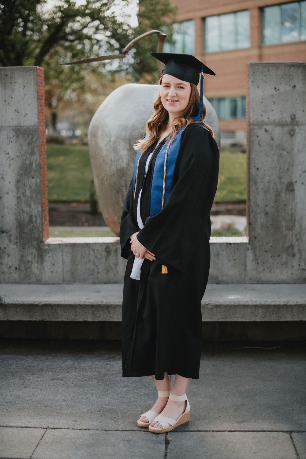 Graduate in black cap and gown with blue sash poses outdoors near concrete pillars on graduation day.