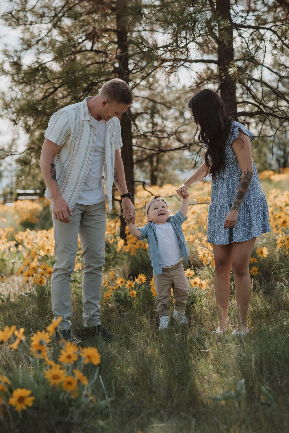 A small child stands in a field of yellow wildflowers with two adults on a sunny afternoon.