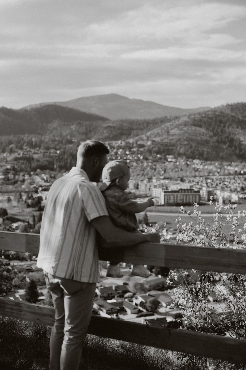 A parent and small child look out over a scenic mountain town vista from an elevated viewpoint in black and white.