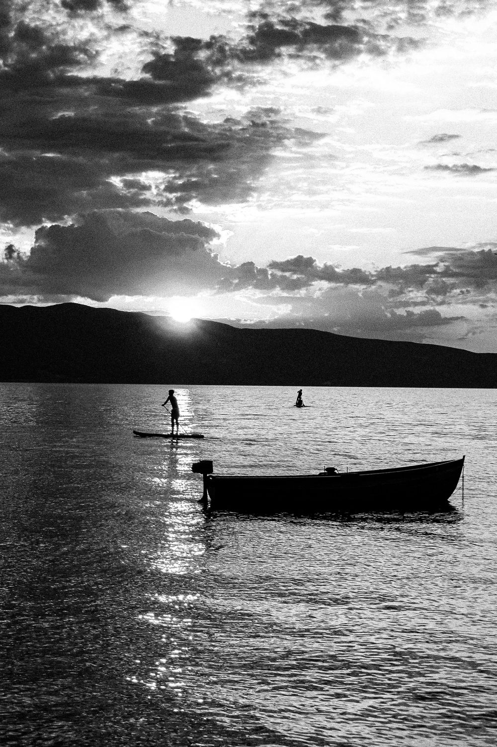 A boy paddling at sunset on Lake Ohrid, a fishing boat in the distance and a anchored boat in the foreground.