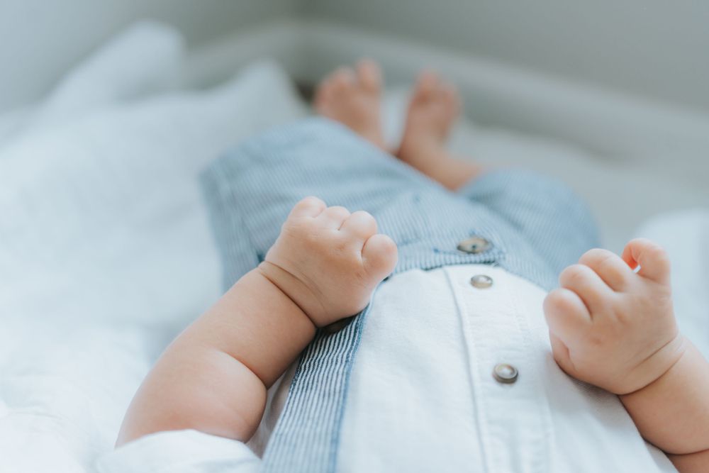 A baby wearing light blue clothing lies on white fabric, with their tiny hands and arms visible in the frame.
