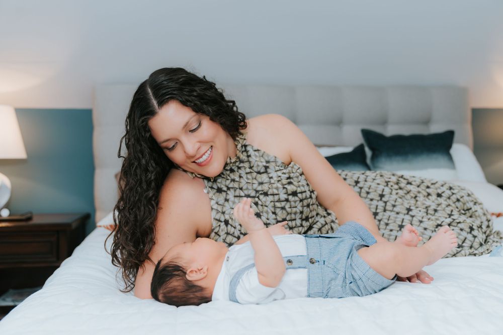Tender moment between parent and baby lying together on white bed wearing floral dress and blue onesie.
