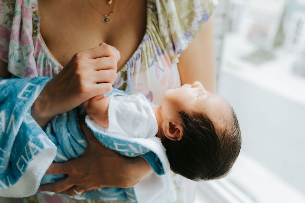 Parent tenderly holds sleeping baby wearing white onesie against soft turquoise fabric in natural light.