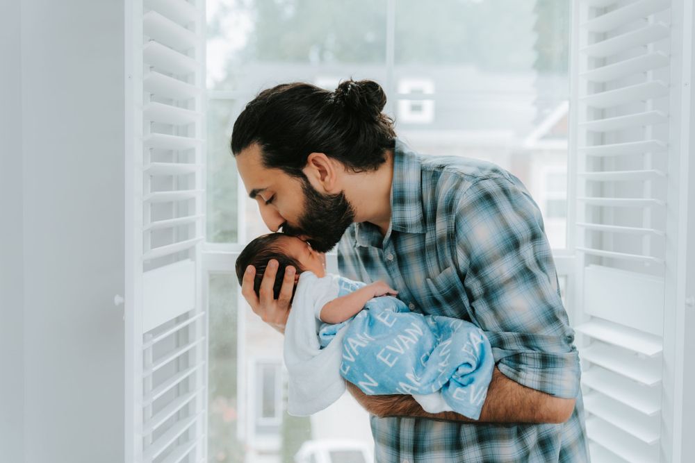 A bearded person in a plaid shirt lovingly holds a swaddled newborn baby near a bright window.
