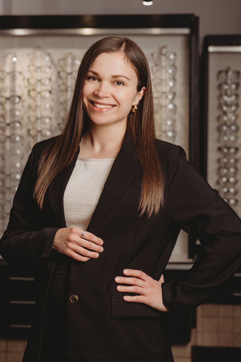 Professional portrait of someone in a black blazer and white top standing in an optical store with eyewear displays.