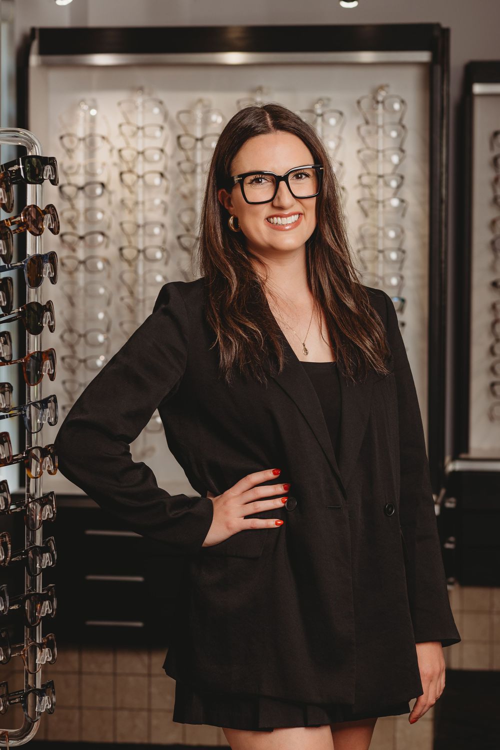 Professional in black blazer and glasses stands in optical store with eyewear display wall in background.