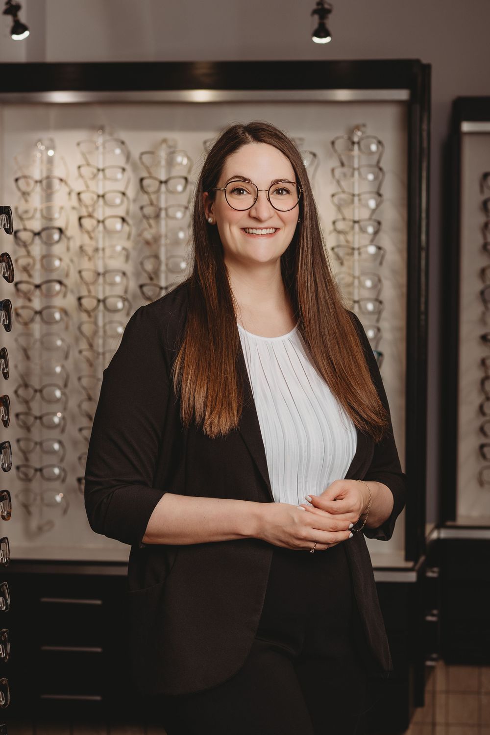 Professional portrait of someone wearing glasses and a black blazer in an optical store with eyewear displays.