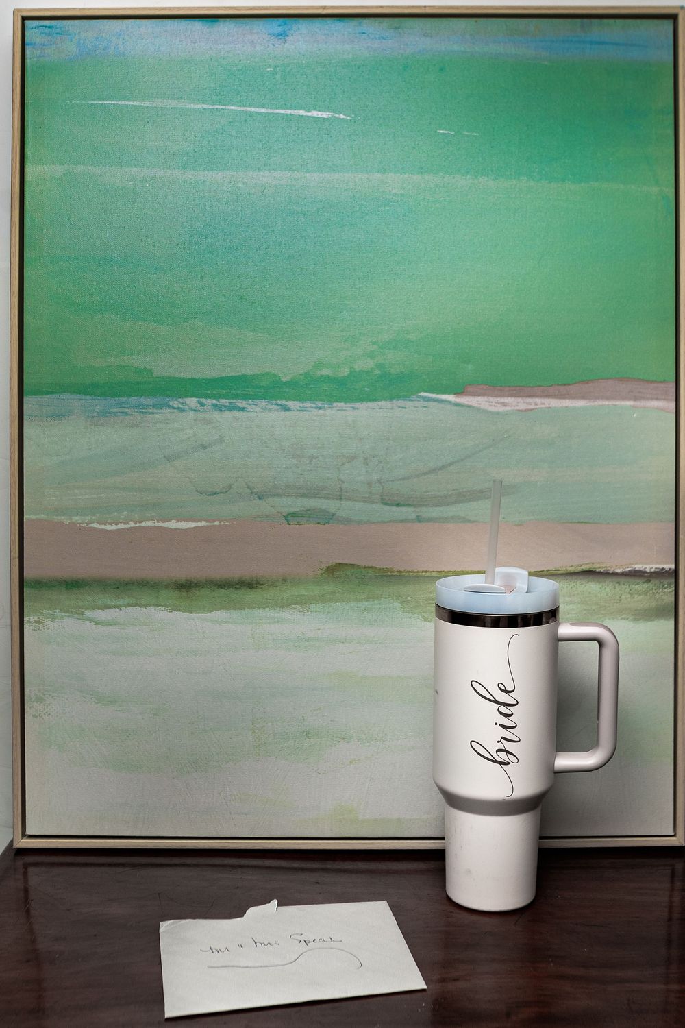 A white travel mug sits on a wooden surface in front of a green and beige abstract landscape painting.