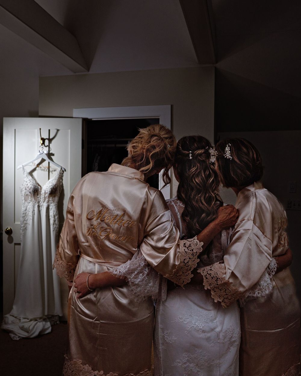 A group of bridesmaids in matching satin robes huddle together before a wedding ceremony in a dimly lit room.