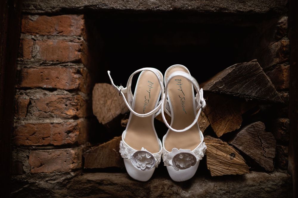 Elegant white bridal shoes with ankle straps displayed against rustic stacked firewood and brick background.