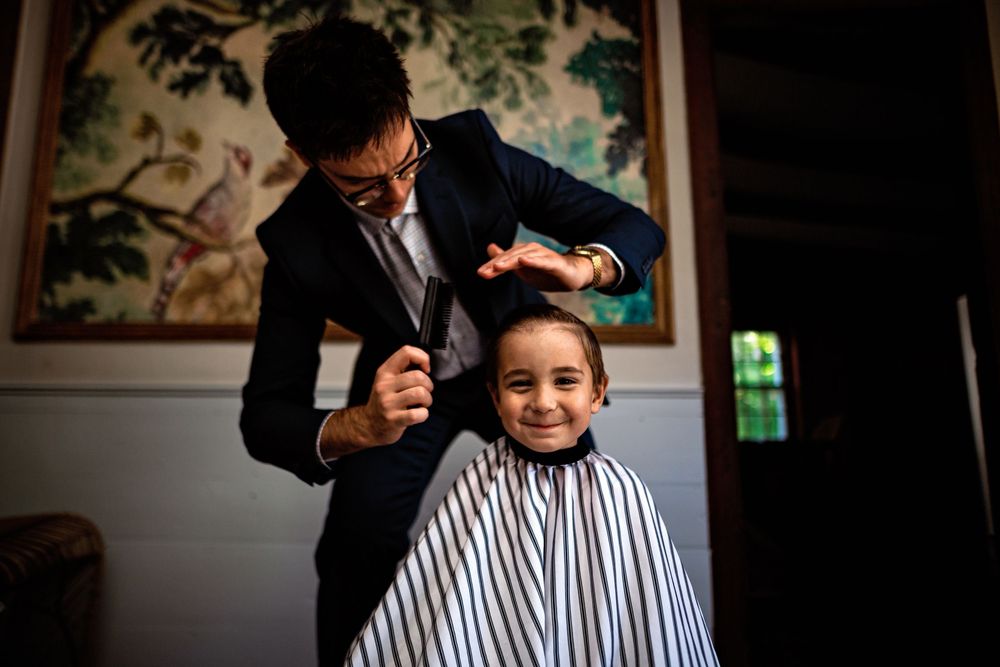 A barber in a black suit styles a client's hair in a striped barber cape at a traditional barbershop.