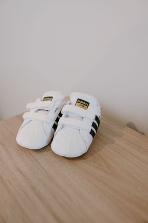 White Adidas Superstar baby shoes with black stripes displayed on a wooden surface.