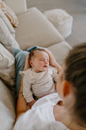 A sleeping baby in a light grey outfit rests on someone's lap on a couch.