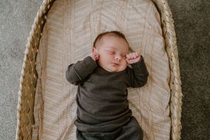 Sleeping baby in dark clothing rests in woven bassinet with geometric pattern bedding.