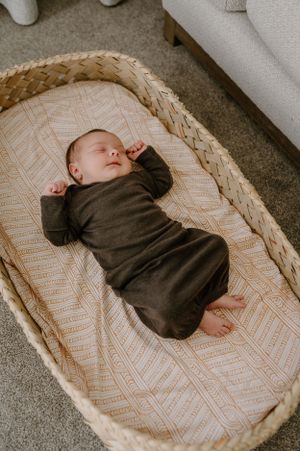 Peaceful sleeping infant in dark outfit lies in woven bassinet on patterned bedding.