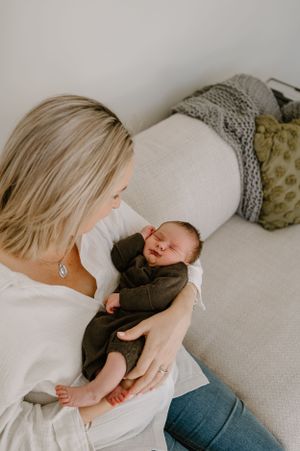 A tender moment captured as a newborn baby in a brown outfit rests peacefully in loving arms on a couch with decorative pillows.