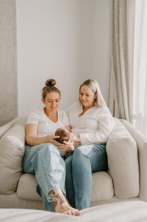 Two people sit on a grey couch in a bright room while holding a small baby.