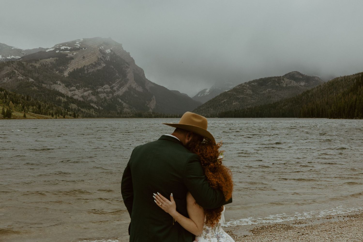 A romantic wedding couple embraces on a dock with autumn foliage and a vibrant red bouquet.
