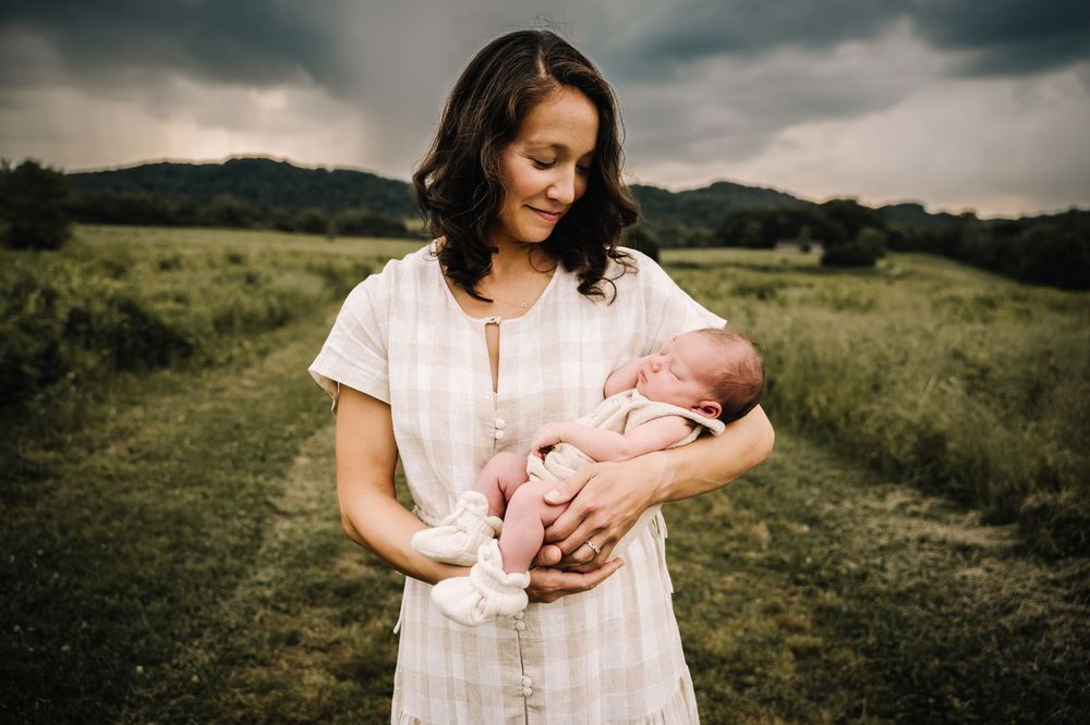 A mother holds her newborn baby in a green field under a cloudy sky. outdoor newborn photos with parents in nashville Tennessee - middle tn