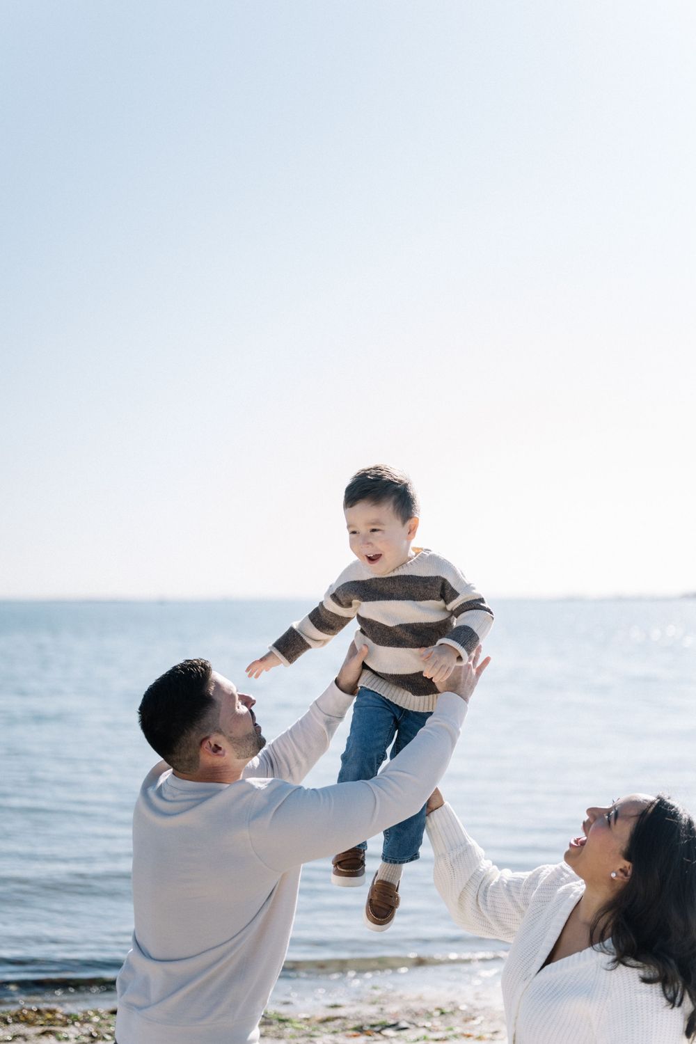 A family plays together on a sunny beach, lifting their child in the air with joy and laughter.
