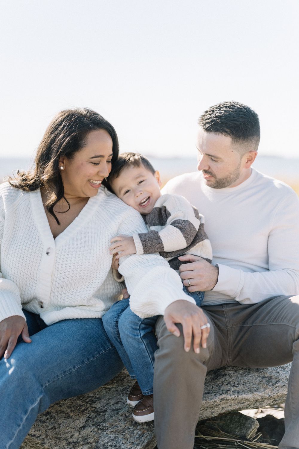 A family in white sweaters and jeans sits together on rocks at the beach sharing joyful moments together.