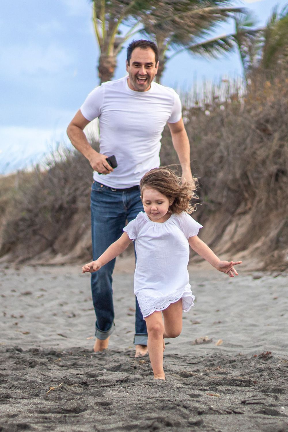 A joyful father chases his laughing young daughter along a sandy beach, both wearing white tops and jeans at Boynton beach maternity.