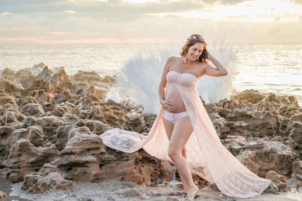 Pregnant woman in flowing pink lace gown poses on rocky beach at sunset with ocean waves crashing behind her Jupiter beach maternity.