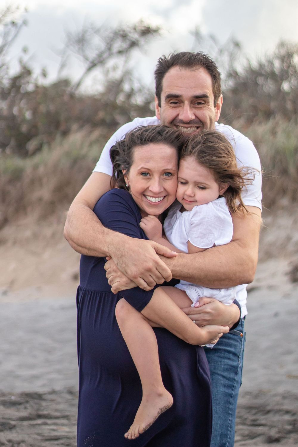Family of three sharing a warm embrace on a sandy beach, parents smiling while holding their young daughter at Boynton beach maternity.