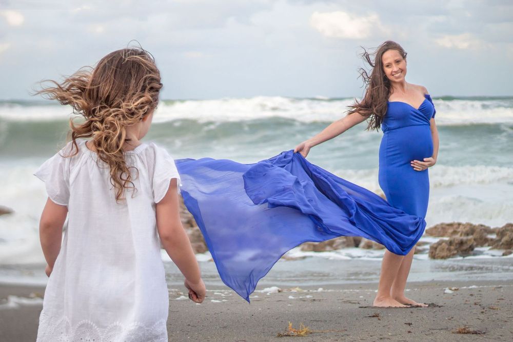 Pregnant woman in blue gown poses on beach with young girl in white dress, ocean waves in background at Boynton beach maternity session.