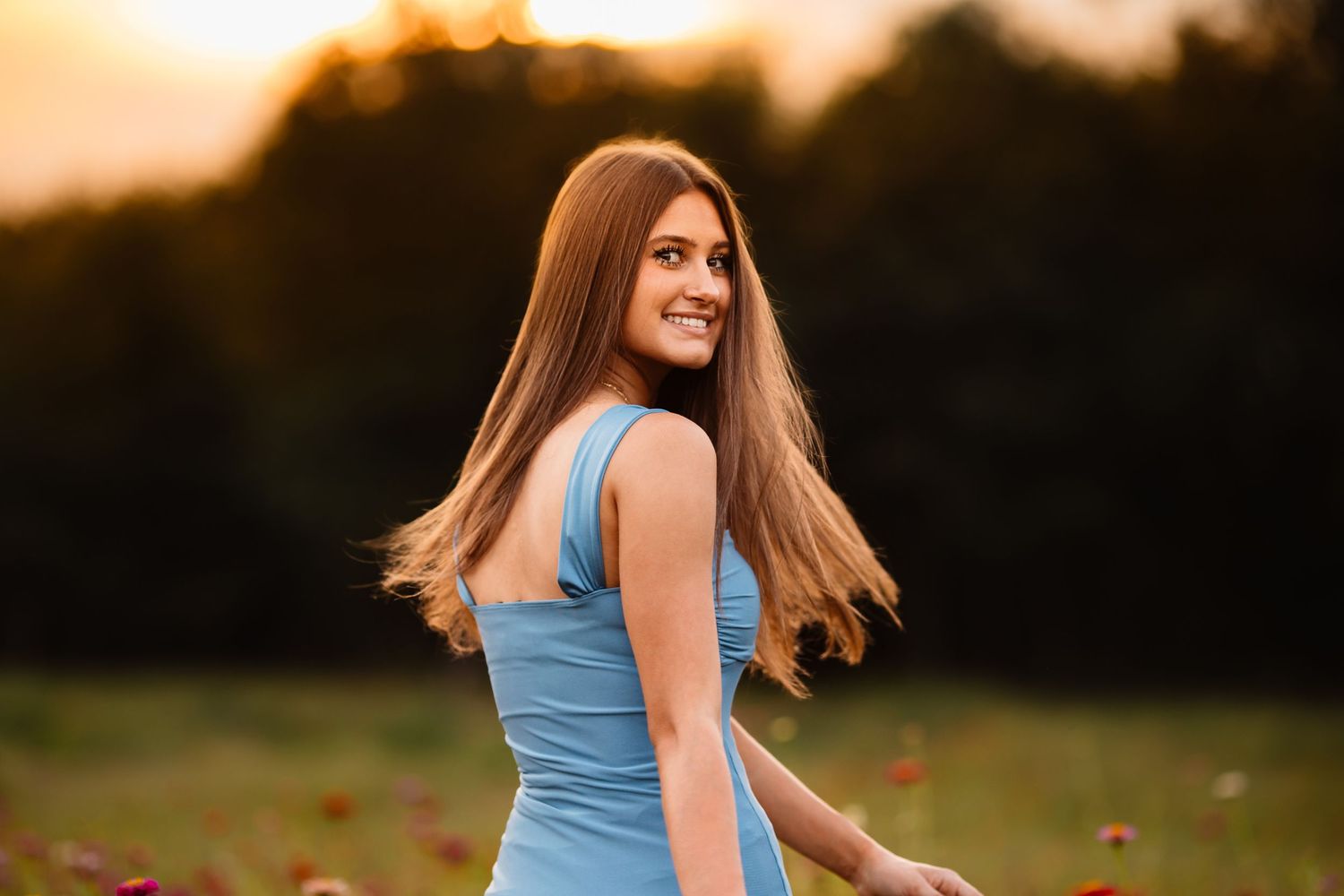 Woman in blue sleeveless dress twirls playfully in a grassy field during golden hour sunset.
