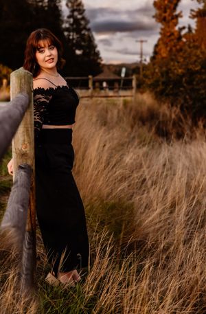 Portrait in black outfit against rustic fence and autumn grass at dusk.