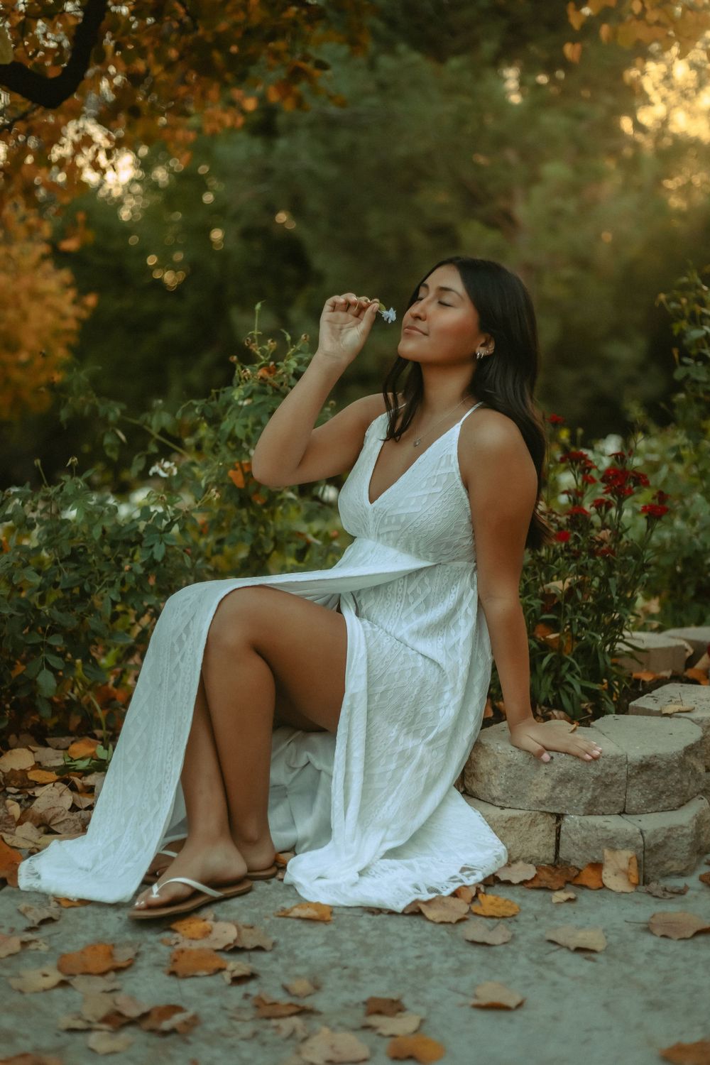 Sweet 16 in a flowing white dress poses gracefully on stone steps surrounded by flowers and garden foliage
