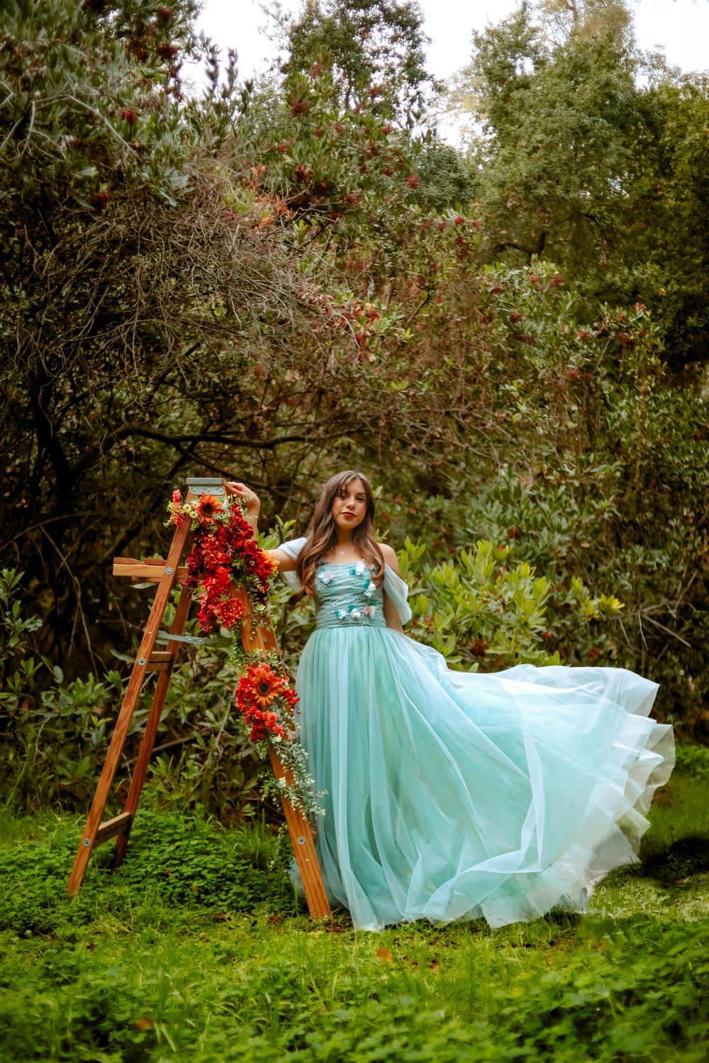 Ethereal turquoise gown flows in an enchanted garden setting with wooden ladder adorned with red floral blooms in San Dimas