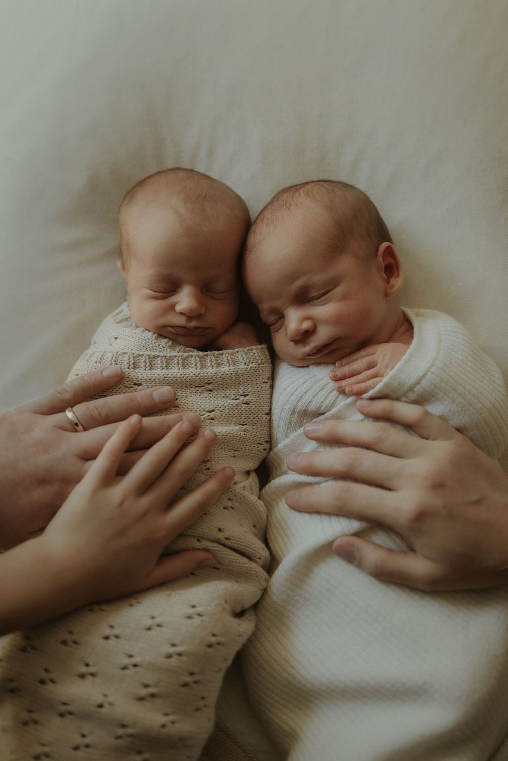 Two sleeping newborn twins in white and patterned outfits lie side by side while gentle hands touch them softly.