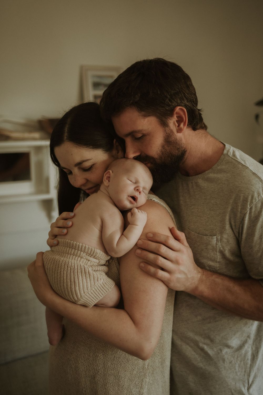 Parents sharing intimate moment with sleeping newborn in warm lighting.