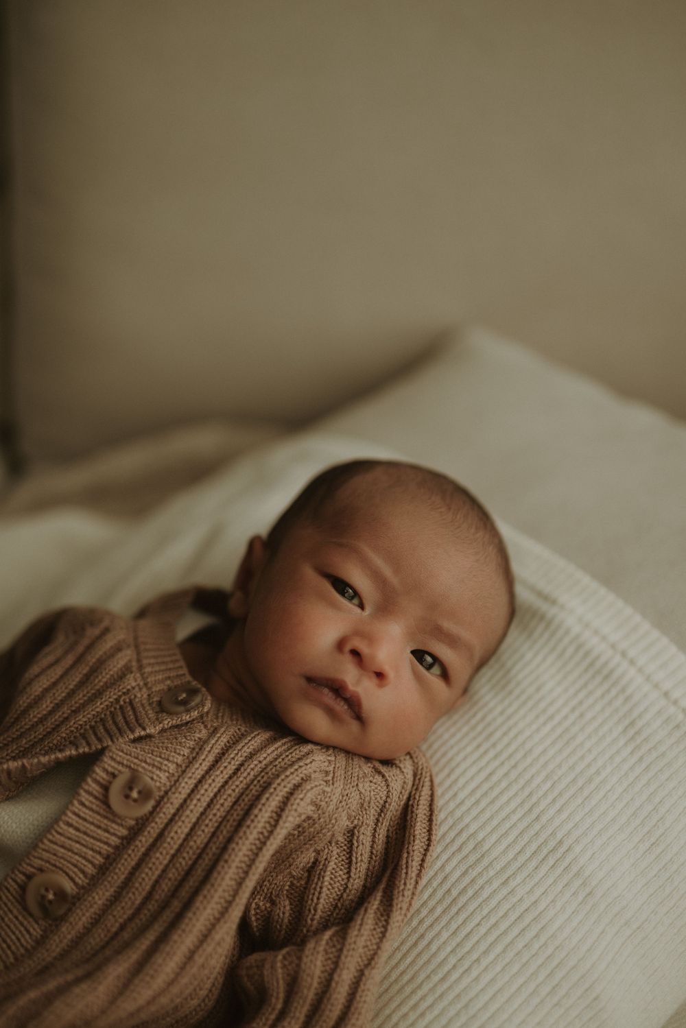 A newborn baby wearing a brown knit sweater lies on white bedding in a warm, moody setting.