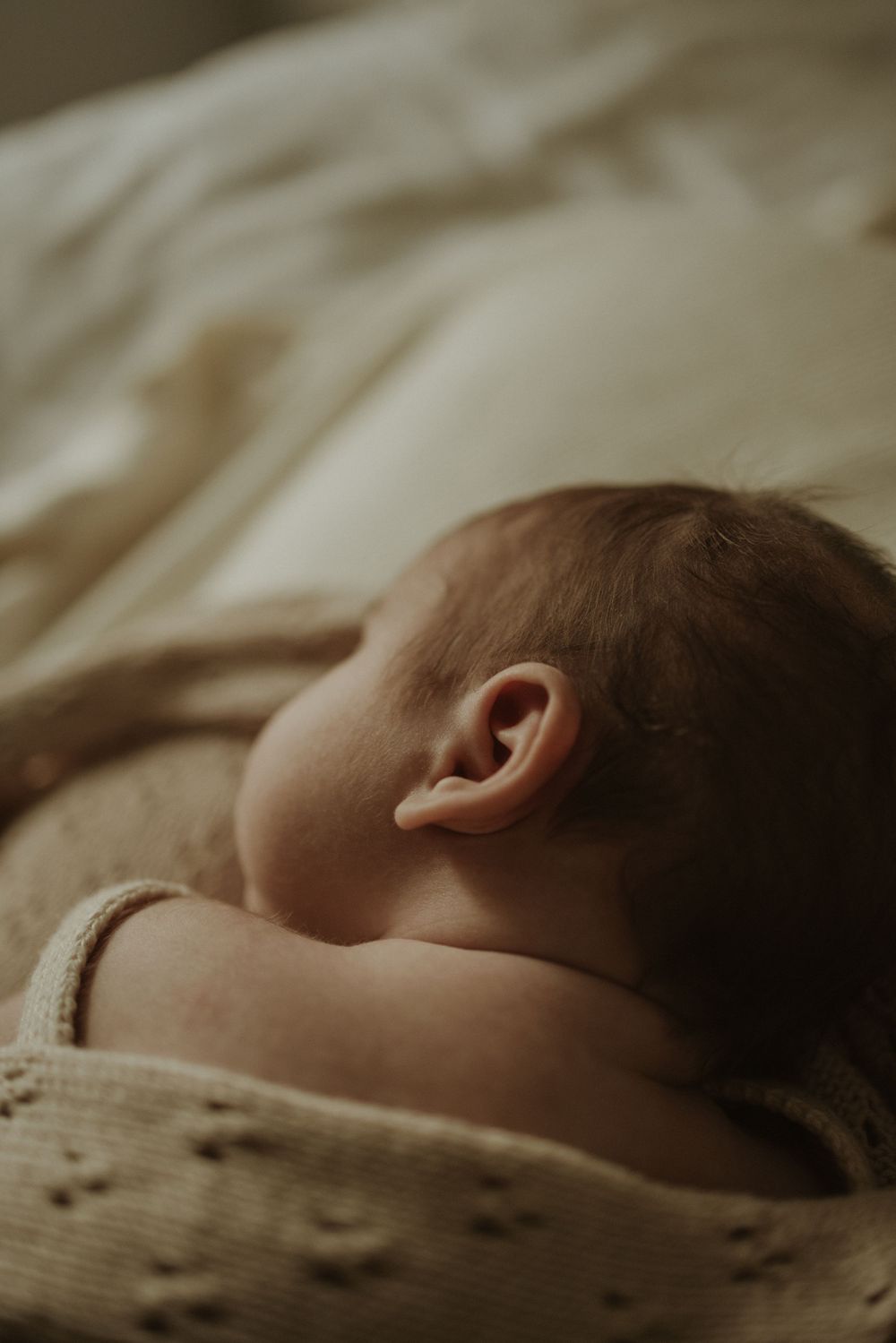 Side profile of sleeping baby showing ear and cheek against textured fabric in warm tones.