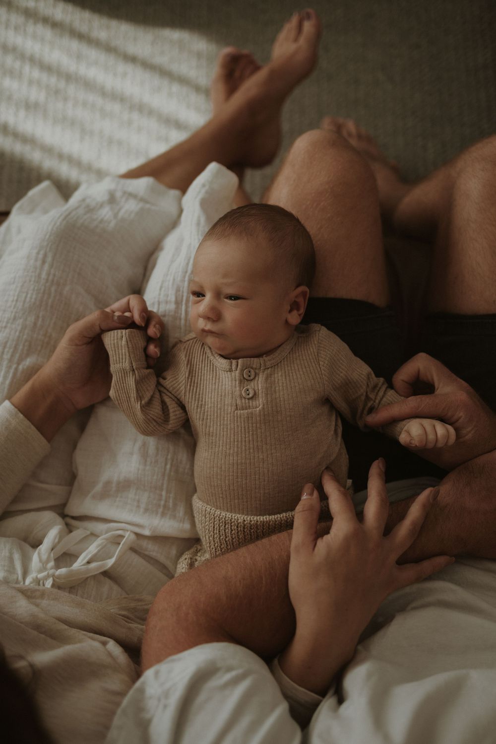 Baby in beige knit outfit lying on white bedding surrounded by caring hands.