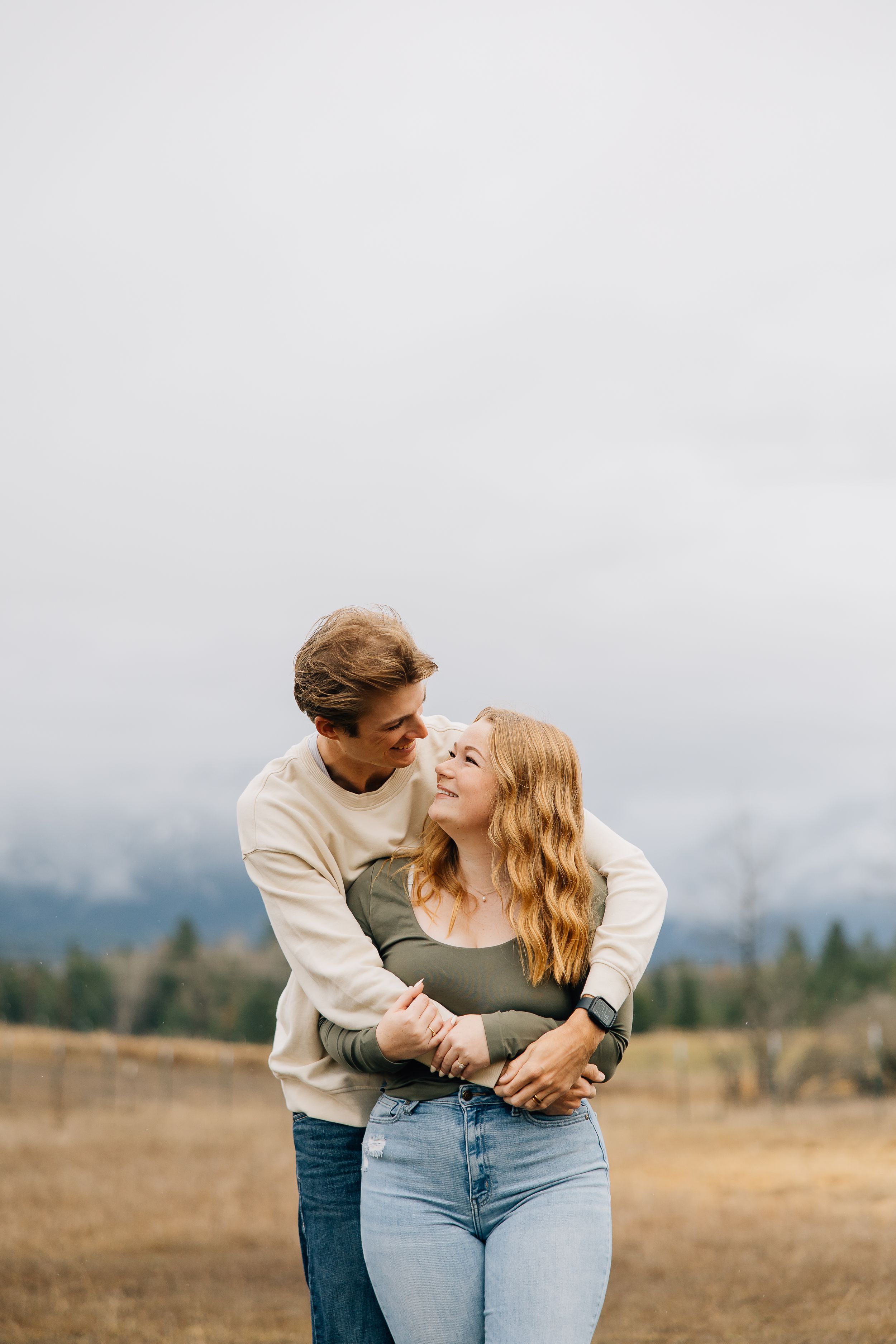 A couple embraces in an intimate moment while standing in a scenic mountain field on an overcast day.