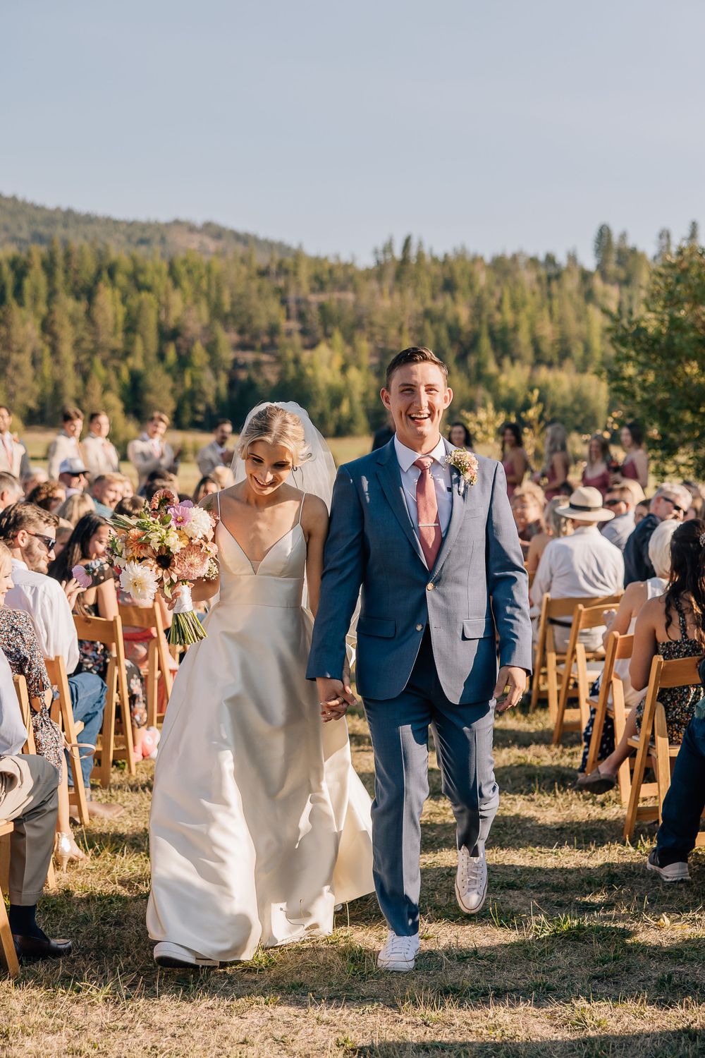 Couple walking down the aisle at an outdoor mountain wedding ceremony with guests seated on wooden chairs.