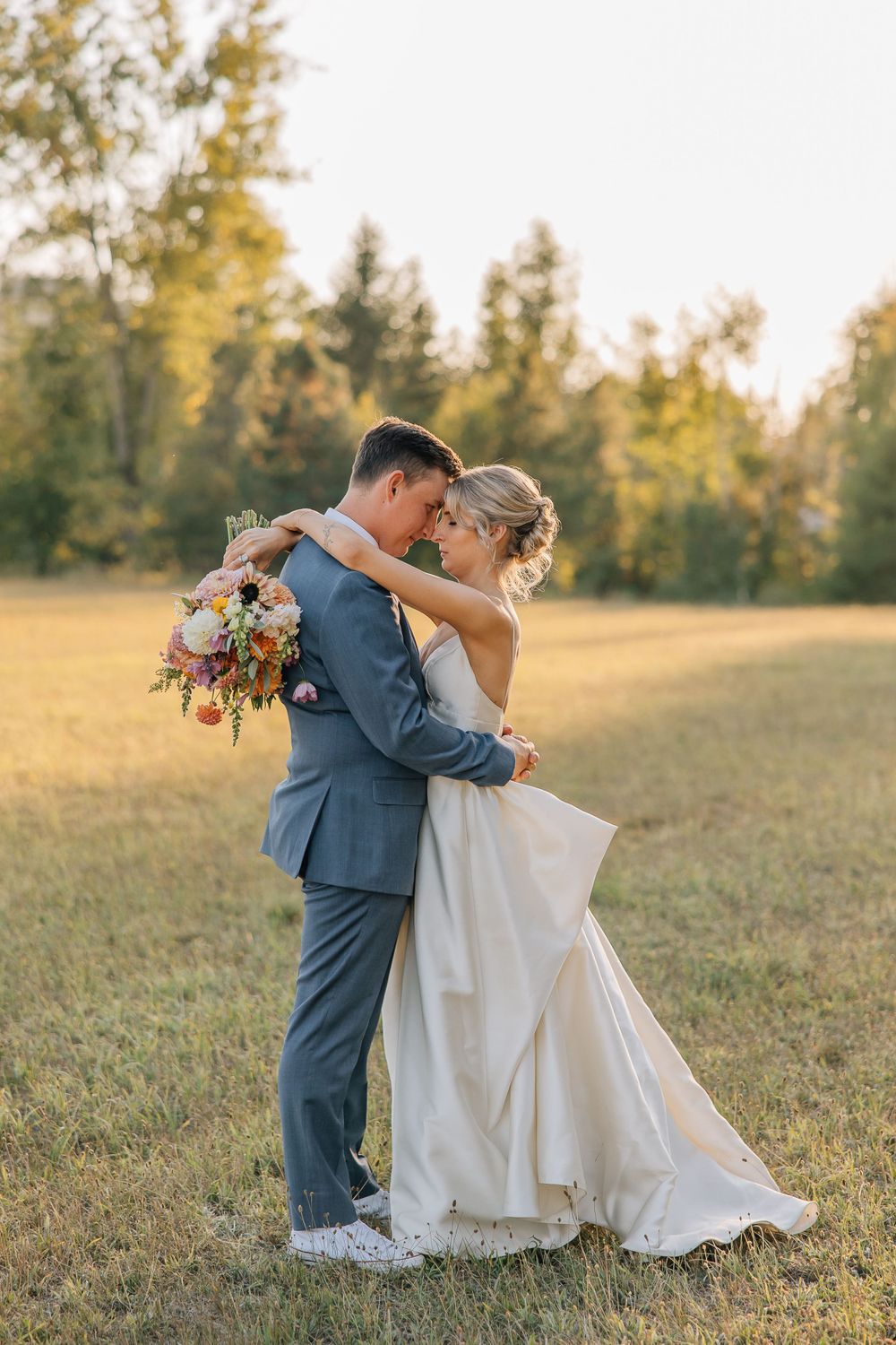 Newlyweds embracing in a golden field during sunset while holding a colorful bouquet.