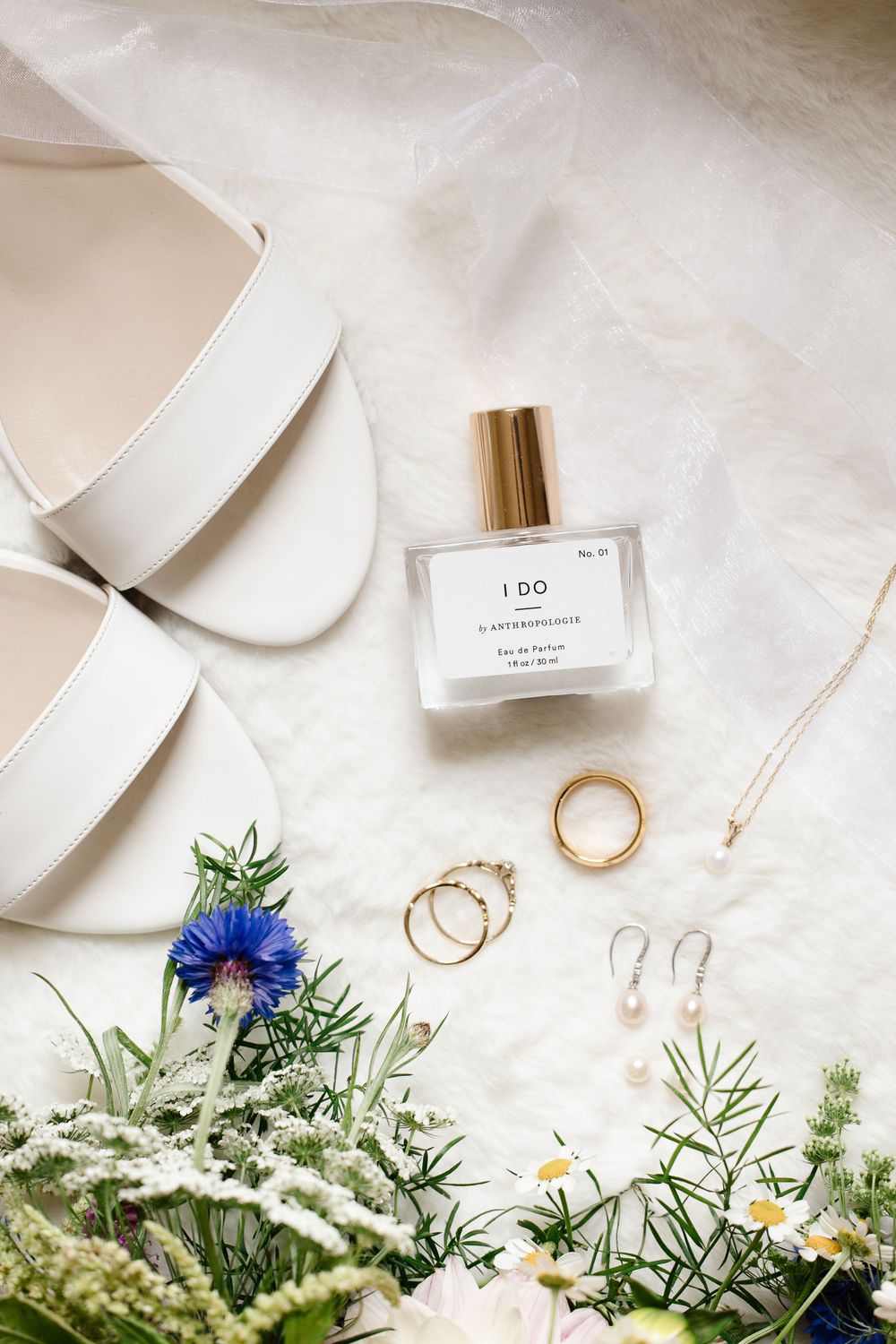 Wedding details flat lay with white shoes, perfume bottle, rings and delicate flowers.