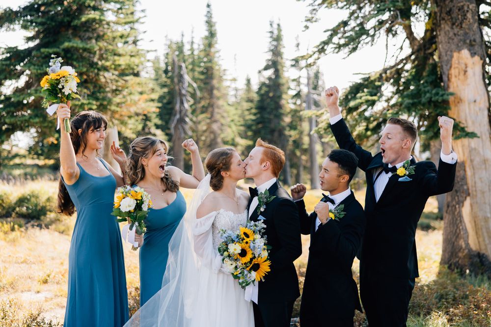 Joyful wedding party celebrates outdoors with raised arms against a backdrop of evergreen trees.